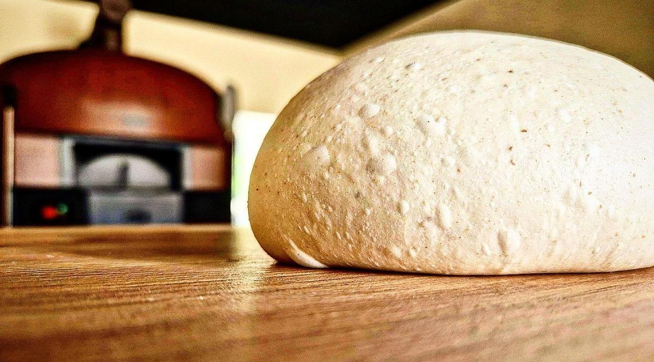 Una pagnotta di impasto sul banco di legno, il forno a legna acceso e sfocato sullo sfondo